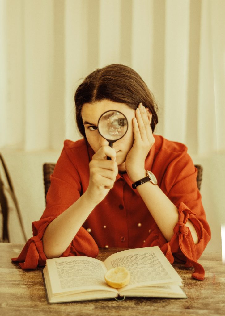 A young woman in a red blouse uses a magnifying glass to examine an open book on a wooden table, with a lemon slice placed on the pages.